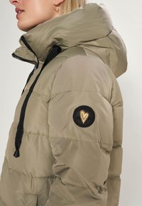 Woman wearing light olive-green puffer jacket with a black circular patch featuring a gold embroidered heart on the upper sleeve.