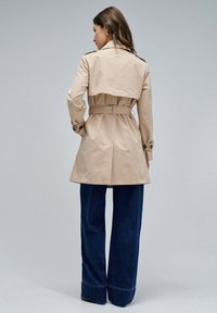 Manteau beige avec une ceinture, épaulettes, et poignets boutonnés. Associé à un jean bleu foncé à jambes larges. Design simple, texture lisse.
