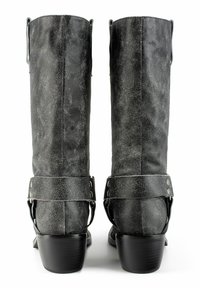 Un par de botas largas de cuero negro desgastado con tacones apilados y correas de arnés vistas desde atrás sobre un fondo blanco.