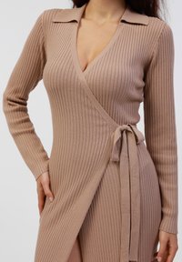 Beige ribbestrikket kjole med lange ermer, V-hals, krage og knyttet midje. Stoffet ser mykt ut med en teksturert finish.