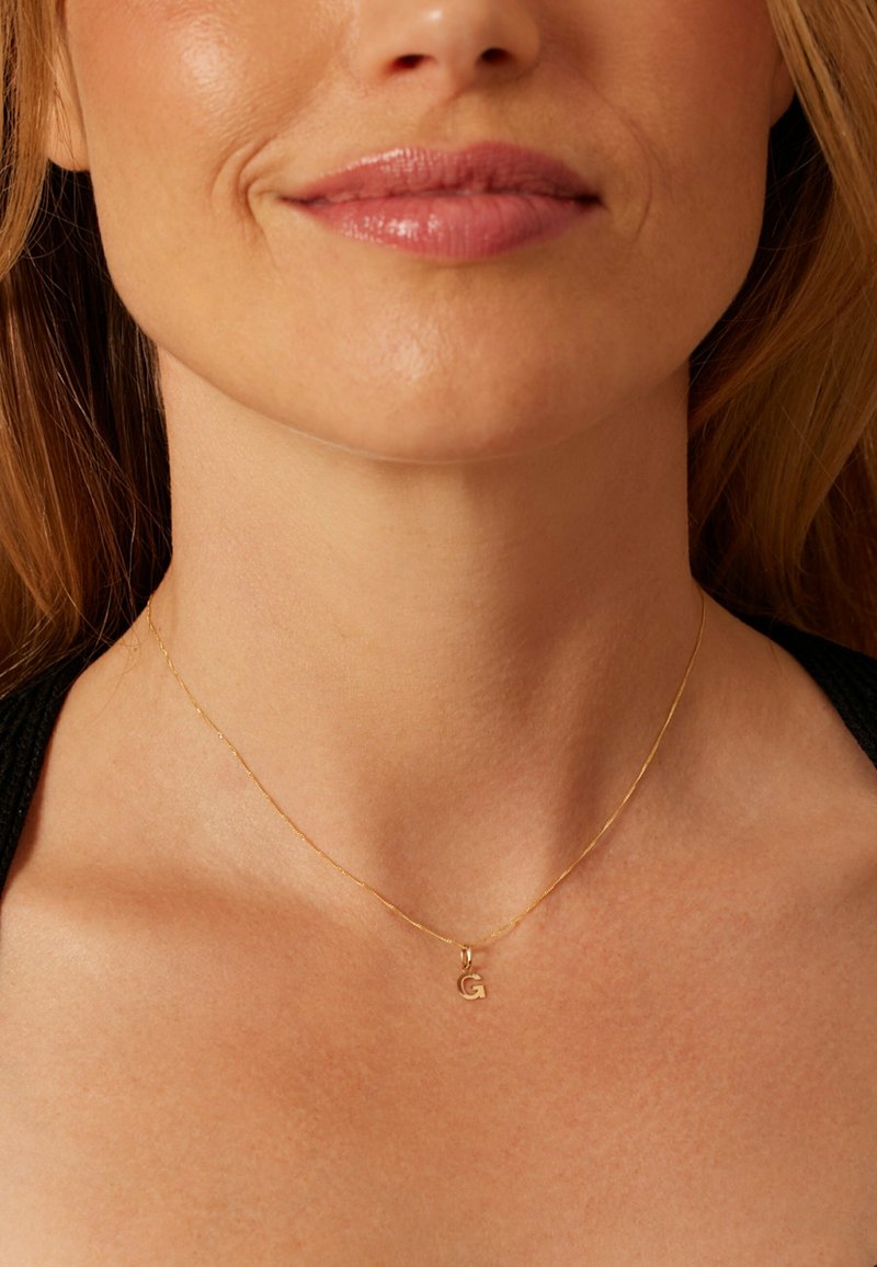 Collier en or avec une chaîne délicate et un petit pendentif "G", porté sur une peau nue. Texture lisse, design fin et style minimaliste.