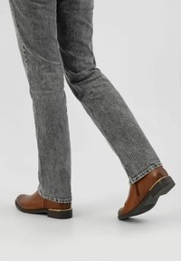 Person, der graue Jeans und braune Leder-Ankle-Boots mit goldenen Akzenten trägt, läuft auf einer hellgrauen Oberfläche.
