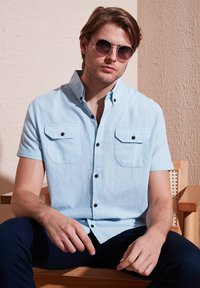 Camisa de botones de manga corta en azul claro con dos bolsillos en el pecho, botones oscuros y un corte relajado, combinada con pantalones navy oscuros y gafas de sol.