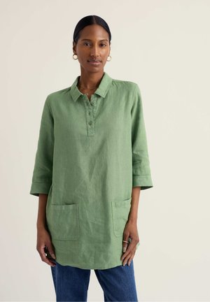 Frau mit dunklem Haar in einem niedrigen Dutt trägt goldene Creolen, ein locker sitzendes grünes Hemd mit Kragen und Taschen vorne sowie eine blaue Jeans, stehend vor einem schlichten Hintergrund.