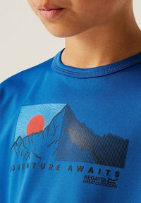 Camiseta azul de manga corta con un gráfico de montaña y sol en rojo, negro y azul, con el texto "La aventura te espera" abajo.