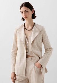Béžový kostkovaný blazer s výstřihem do V, spárovaný s odpovídajícím topem s nízkým výstřihem a ozdobnými knoflíky a elegantními kalhotami.