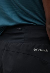 Κοντινό πλάνο ατόμου που φοράει μαύρο παντελόνι Columbia με τσέπη με φερμουάρ και μπλούζα με υφή σε τυρκουάζ χρώμα.