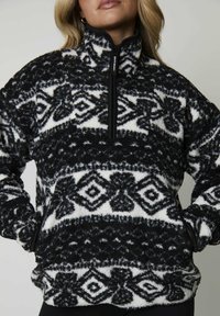 Pullover in pile bianco e nero con motivo a disegni, colletto alto, chiusura con zip frontale e tasche laterali. Presenta design geometrici e una superficie testurizzata.
