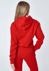 Felpa rossa con cappuccio, tasca frontale e cordino regolabile. Realizzata in tessuto morbido, presenta polsini e vita a coste.