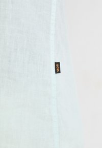 Világoskék vászon ing, finom textúrával, fekete címkével, rajta narancssárga "BOSS" felirattal a varrat közelében. Egyszerű dizájn, egyenes szabás.
