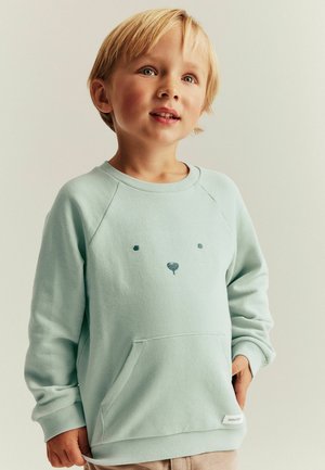 Młody chłopiec o blond włosach, ubrany w jasnoniebieską bluzę z prostym wzorem twarzy misia, stojący na jednolitym tle.
