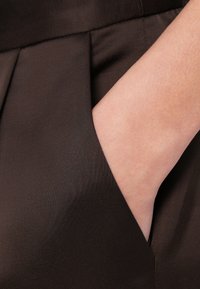 Tissu brun texturé avec un motif plissé, montrant une main s'insérant dans une poche latérale, mettant en valeur la forme et l'ouverture de la poche.