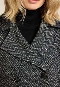 Houndstooth-jas in zwart en wit met grijze accenten, versierd met pailletten. Heeft een grote kraag en dubbele knopen.