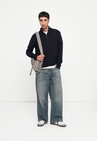 Marineblauer Pullover mit Reißverschluss am Hals, kombiniert mit locker sitzenden, ausgebleichten blauen Jeans. Graue Umhängetasche mit mehreren Fächern. Weiße Sneaker.
