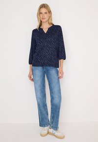 Blouse bleu marine avec un motif en chevron, décolleté en V et manches trois-quarts, associée à un jean droit bleu clair et des baskets blanches.