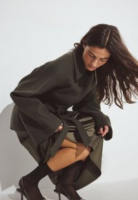 Olivnozelena volnena bunda z širokimi rokavi in pasom, oblečena čez satenasto krilo. Prosojne črne nogavice in odprti petki dopolnjujejo videz.