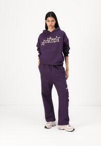 Hanorac mov cu text grafic și pantaloni de trening mov asortați. Prezintă o croială lejeră, o textură moale și accente de crem pe hanorac.