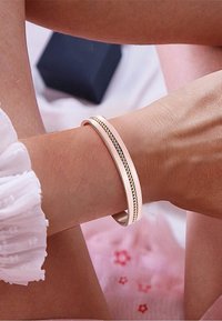 Bracciale rigido in tono oro con superficie liscia e dettaglio a corda intrecciata al centro. Indossato su un polso con uno sfondo morbido e testurizzato.