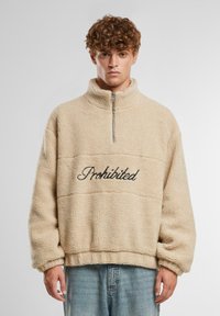 Pullover in pile beige con colletto alto, mezza cerniera e ricamo nero "Proibito" sul davanti. Presenta una vestibilità comoda e un tessuto testurizzato.