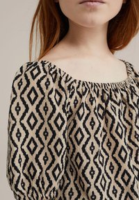 Béžový top s černým diamantovým vzorem, se čtvercovým výstřihem a nařasenými rukávy, vyrobený z texturované látky s mírným balónovým efektem.
