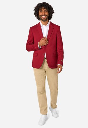 Glimlachende man met krullend haar, gekleed in een rode blazer, wit overhemd, beige broek en witte sneakers, staand tegen een effen achtergrond.