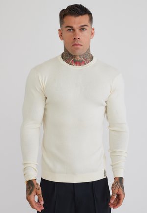 Mann mit Hals- und Handtattoos, der einen eng anliegenden cremefarbenen, gerippten Langarmpullover und schwarze Hosen trägt und vor einem schlichten Hintergrund steht.