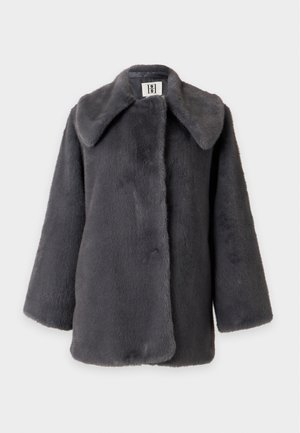 Vrouw draagt een donkergrijze oversized nepbontjas, zwarte coltrui, zwarte broek en zwarte open-teen hakken, staand tegen een effen lichte achtergrond.