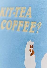 Svetlo modrá tkanina s krémovým vyšívaním a textom "KIT-TEA COFFEE?" a bielou rukou s hnedými otlačkami labiek.