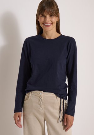 Femme portant une chemise bleu marine à manches longues avec un pantalon beige, souriante et debout devant un fond clair uni.