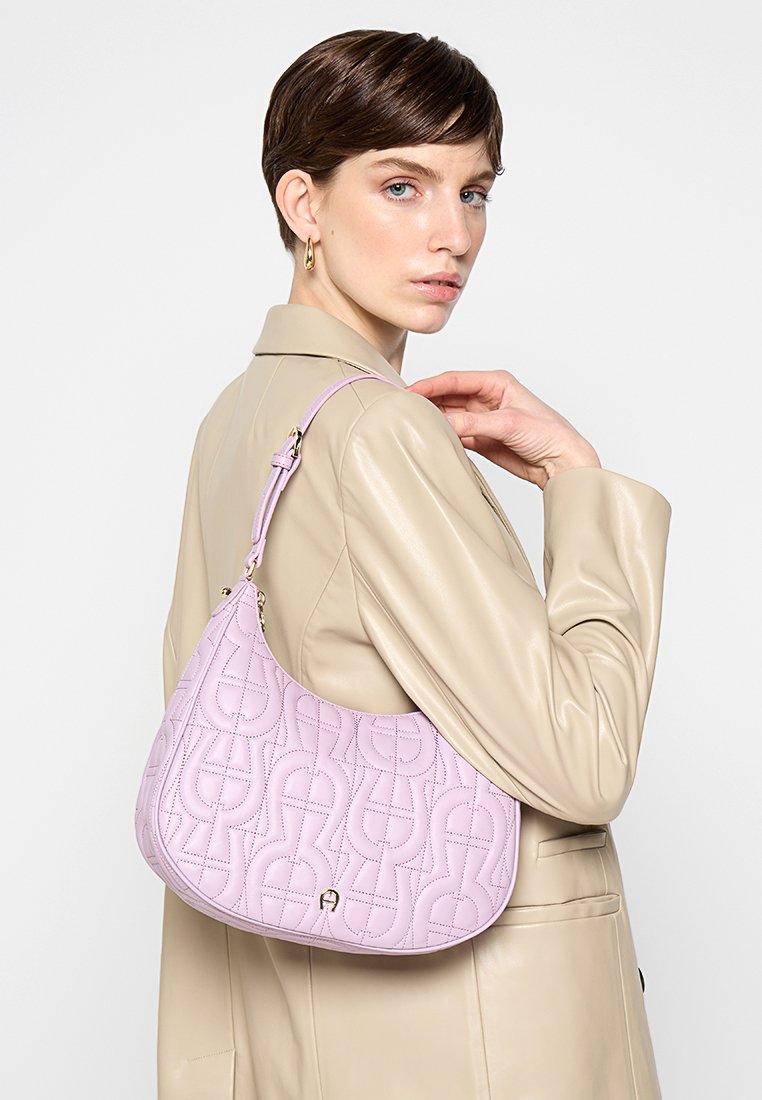 Frau in beiger Lederjacke, die eine helllila, gequiltete Schultertasche mit geometrischen Mustern über der Schulter trägt.