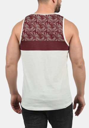 Mann trägt ein ärmelloses weißes Tanktop mit einem weinroten blumigen Muster auf dem oberen Rückenbereich und einem einfarbigen weinroten Streifen darunter.