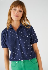 Maglietta polo blu navy con pois bianchi, dotata di colletto e chiusura con bottoni, abbinata a pantaloni verdi. Tessuto morbido con maniche corte.