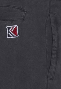 Mörkgrått tyg med en broderad röd, vit och blå geometrisk logotyp nära en ficka med klaff och ett vertikalt slitsöppning.