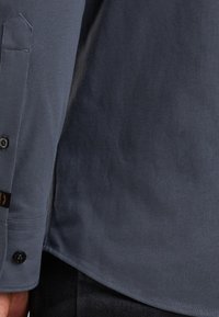 Primer plano de una persona que lleva una camisa abotonada de color gris oscuro y pantalones negros, mostrando el puño de la camisa, los botones y la textura de la tela.