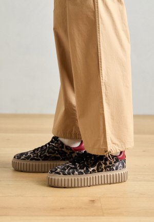 Person iført beige bukser med vide ben og leopardprint sneakers med tykke beige såler, stående på et trægulv.