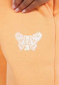 Peachfarvede sweatpants i et blødt stof har et hvidt blomster-butterfly logo med tekst. Side-lomme synlig med kontrasterende syning.
