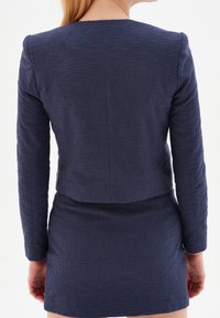 Femme portant un blazer à manches longues bleu marine texturé et une jupe assortie, vue de dos sur un fond blanc.