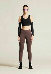 Schwarzes Croptop, lange schwarze Armschläuche und braune Leggings. Als Zubehör gibt es reflektierende Sonnenbrillen und schwarze Sportschuhe. Glatte Texturen.