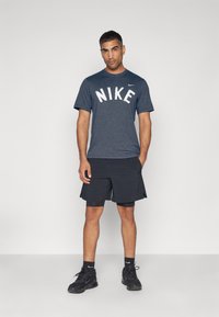 Vīriešu sporta apģērbs, kas sastāv no tumši pelēka Nike T-krekli ar balto logotipu, melnām šortiem un melnām sporta kurpēm. Gluda auduma un ikdienišķs dizains.