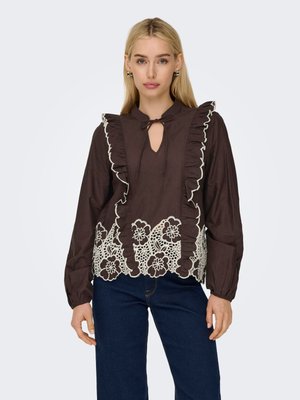 Donna bionda che indossa una camicetta marrone scuro con rifinitura arricciata, ricamo floreale in pizzo all'orlo e jeans blu scuro, in piedi davanti a uno sfondo semplice.