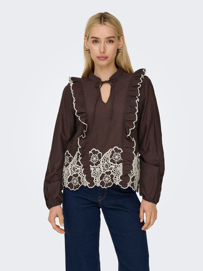Femme blonde portant un chemisier brun foncé avec bordure à volants, broderie florale en dentelle à l'ourlet, et jean bleu foncé, debout devant un fond uni.