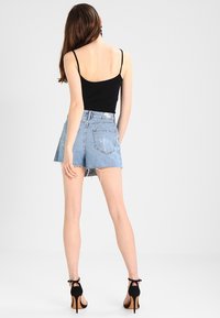 Top negro sin mangas y shorts de mezclilla azul claro con detalles desgastados. Llevados con sandalias negras de tacón alto, vistos desde atrás.