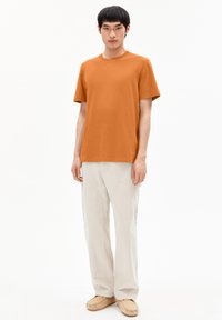 Orange T-Shirt mit kurzen Ärmeln, Rundhalsausschnitt, kombiniert mit hellbeigen Hosen und lässigen Slippern. Schlichte Textur, keine Muster.