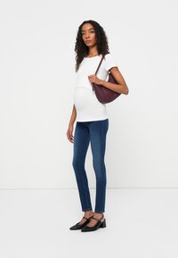 Modelo embarazada con una blusa blanca de manga corta, jeans ajustados azules y zapatos de tacón negros, sosteniendo un bolso burdeos.