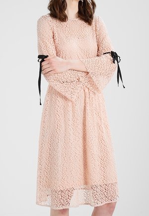 Frau trägt ein knielanges, langärmliges pfirsichfarbenes Spitzenkleid mit schwarzen Schleifen an den Ärmeln, die Arme verschränkt.