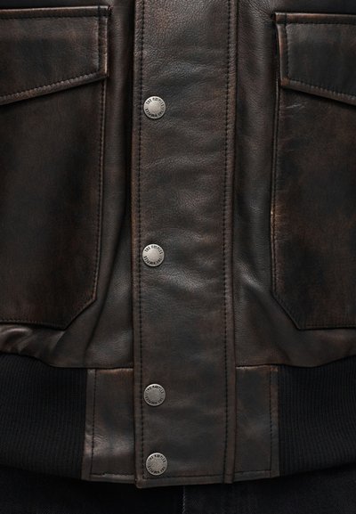 Καφέ δερμάτινο μπουφάν με λεία υφή, μεταλλικά κουμπώματα και δύο μπροστινές τσέπες, με ριγέ μαύρο τελείωμα στο κάτω μέρος.