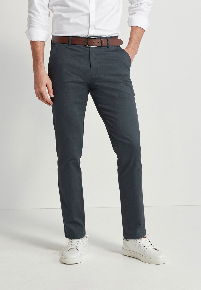 Pantaloni grigi scuri su misura con un tessuto liscio, abbinati a una cintura in pelle marrone e sneaker bianche. Presentano un design a gamba dritta.