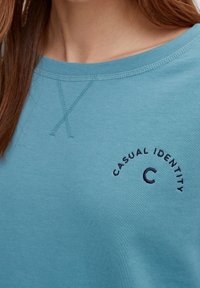 Sweatshirt bleu clair avec un col rond, portant un texte brodé « CASUAL IDENTITY » en bleu marine. Texture douce et lisse.