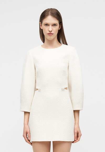 Γυναίκα με ίσια καστανά μαλλιά που φοράει λευκό κοντό φόρεμα με μακριά μανίκια, υφασμάτινη υφή, μικρές ανοίγματα και διακοσμητικά φιόγκους στη μέση.