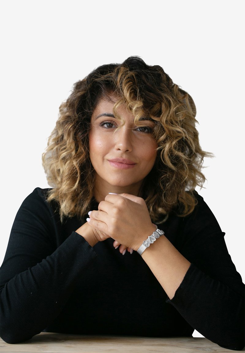 Bracelet en argent avec des accents de papillon, porté au poignet. Le sujet a les cheveux bouclés, porte un haut noir à manches longues, les mains jointes.